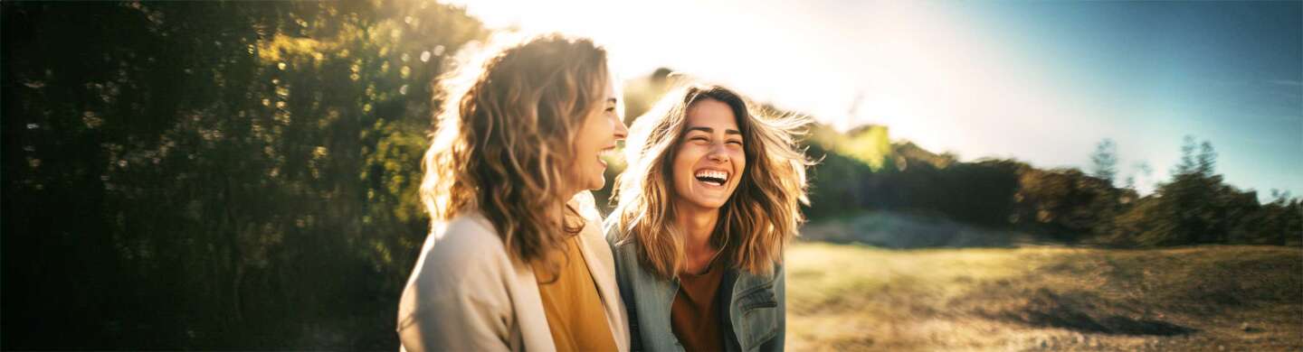 Zwei Frauen sitzen lachend vor einer Waldlandschaft.