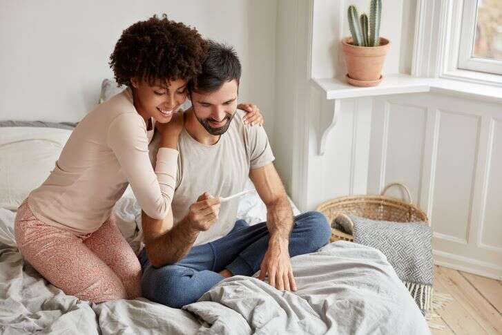 Ein Mann und eine Frau sitzen auf einem Bett und blicken erfreut auf einen Schwangerschaftstest.